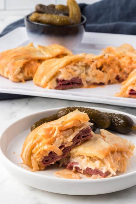 Pickles and a puff pastry corned beef sandwich on a plate in the foreground with more slices behind it.