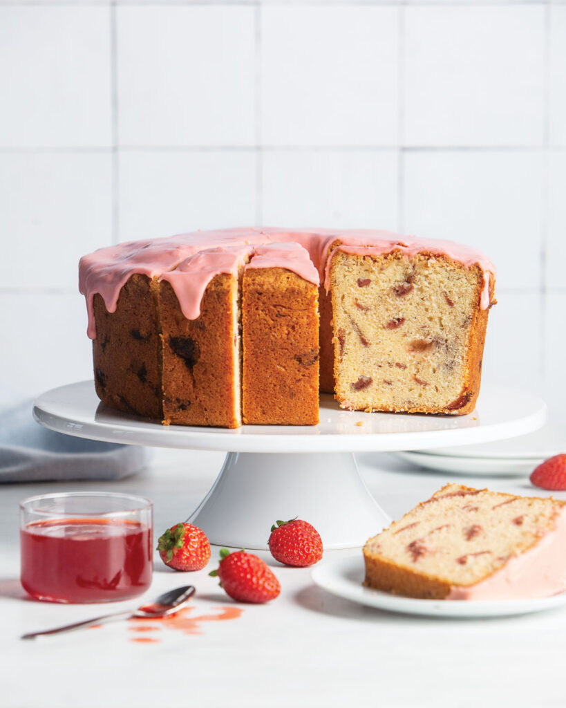 Roasted Strawberry Pound Cake on white cake stand with one slice of cake on white plate and jar of strawberry jam with spoon and whole strawberries