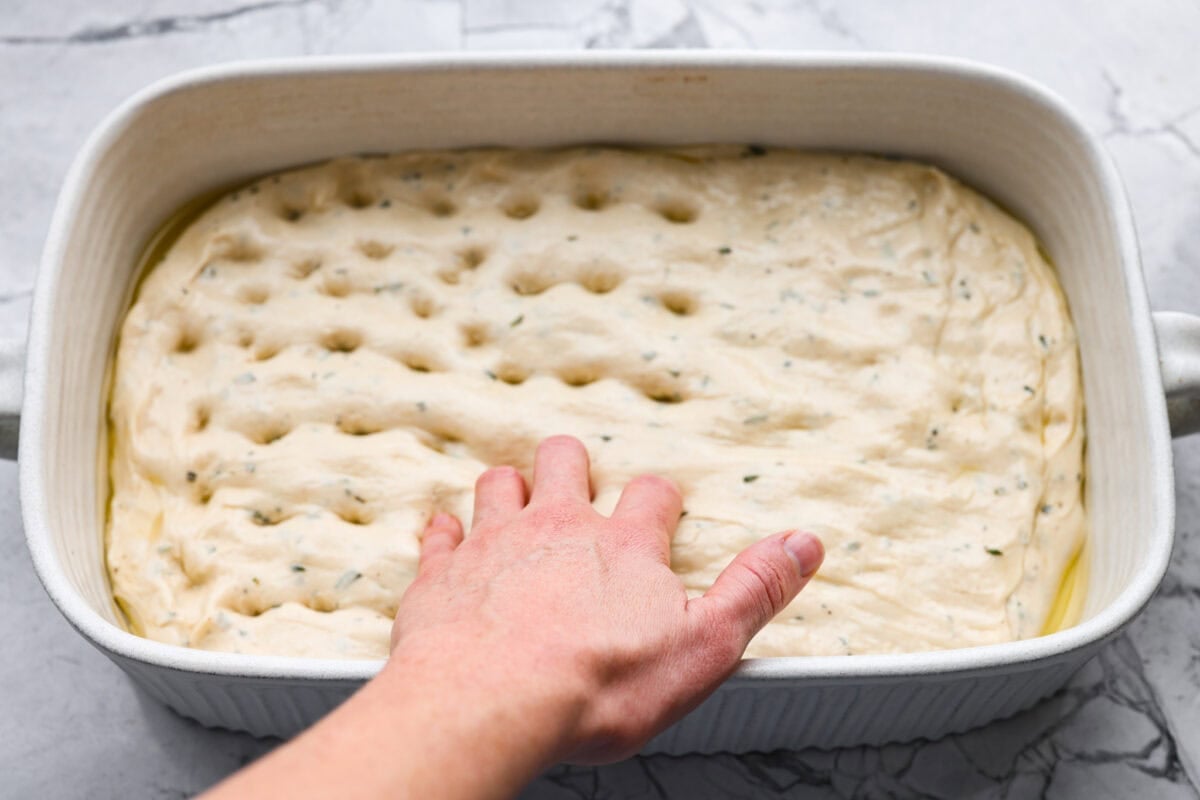 Someone using their fingers to poke holes into the focaccia dough in a baking dish. 
