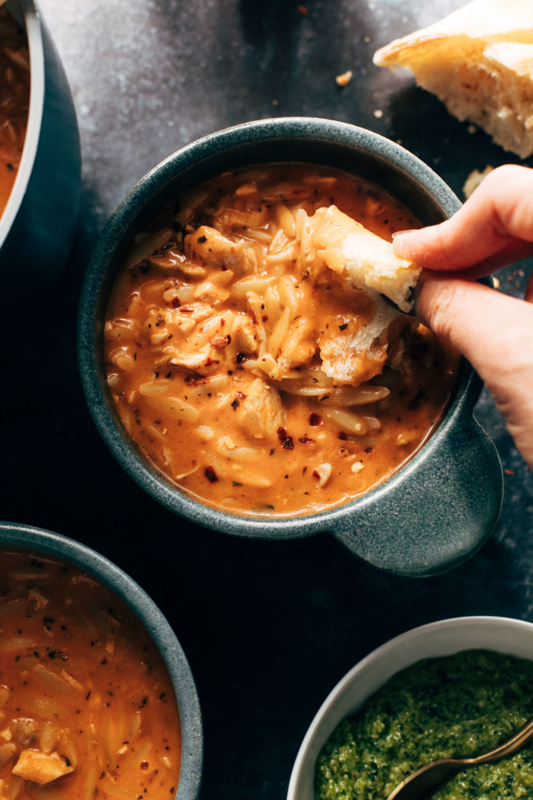 Chicken and orzo tomato soup in a bowl with bread being dipped in.