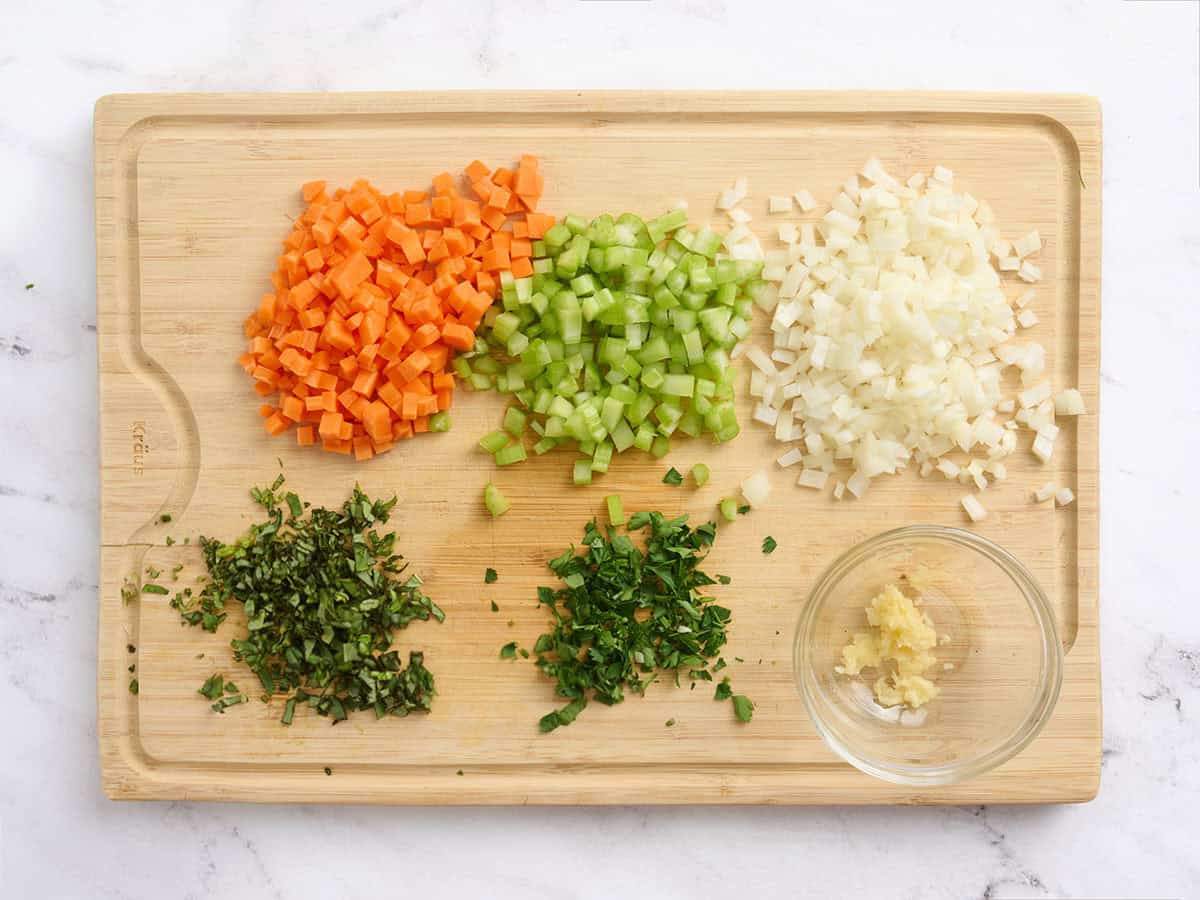 Diced carrots, onion, celery and minced herbs and garlic on a wooden cutting board.