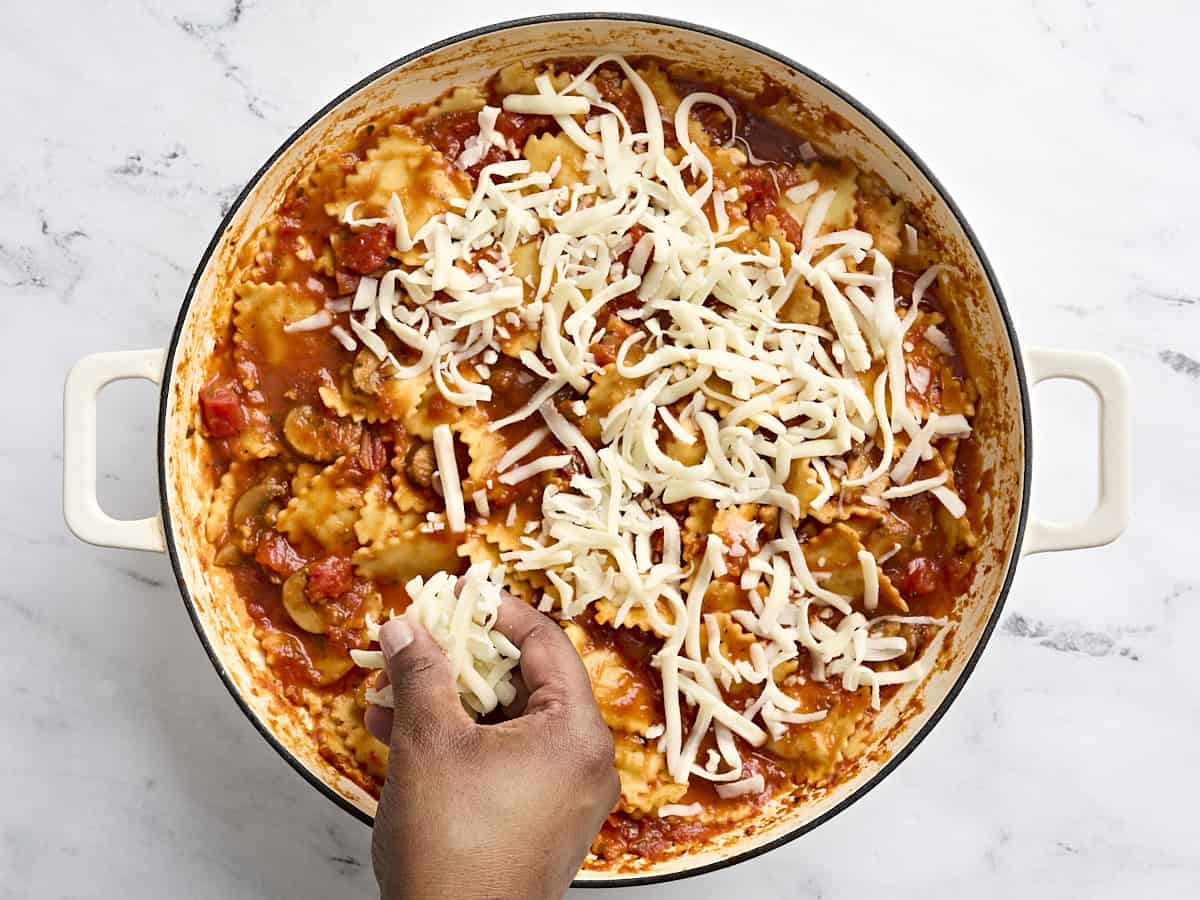 A hand sprinkling shredded cheese over cheese ravioli in a skillet.