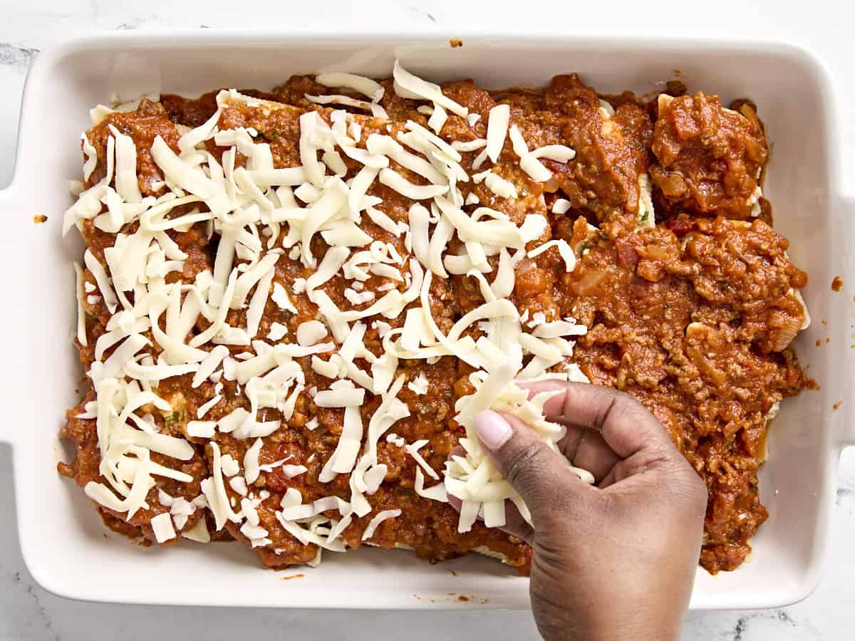 Shredded cheese being added on top of stuffed shells in a casserole dish.