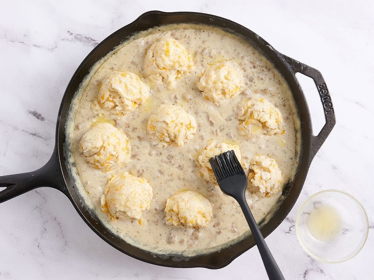 A pastry brush brushing butter over Finished biscuits and gravy bake in a skillet before baking.