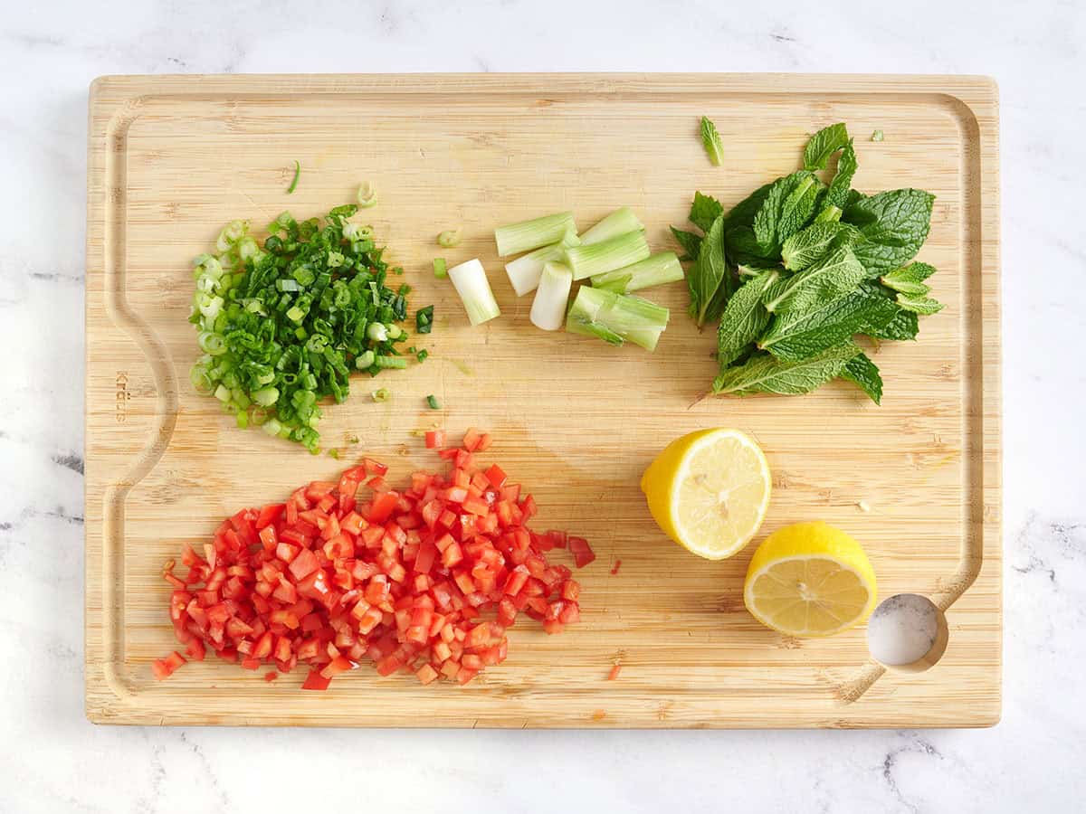 Diced tomatoes, a lemon sliced in half, minced green onion and mint on a chopped board.