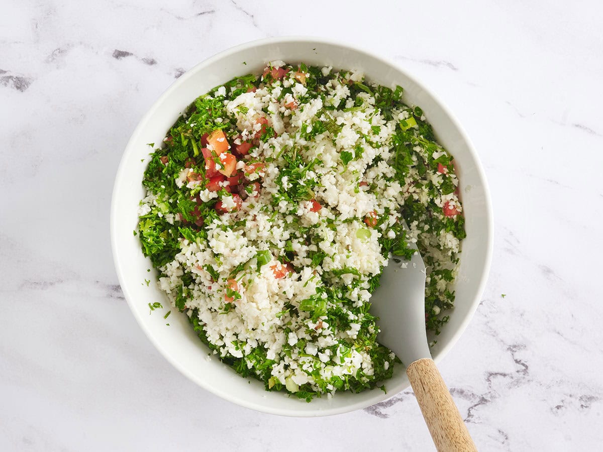 A spatula folding a cauliflower tabbouleh in a bowl.