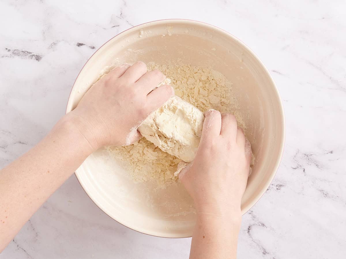 Pie dough ingredients being combined in a mixing bowl by two hands.