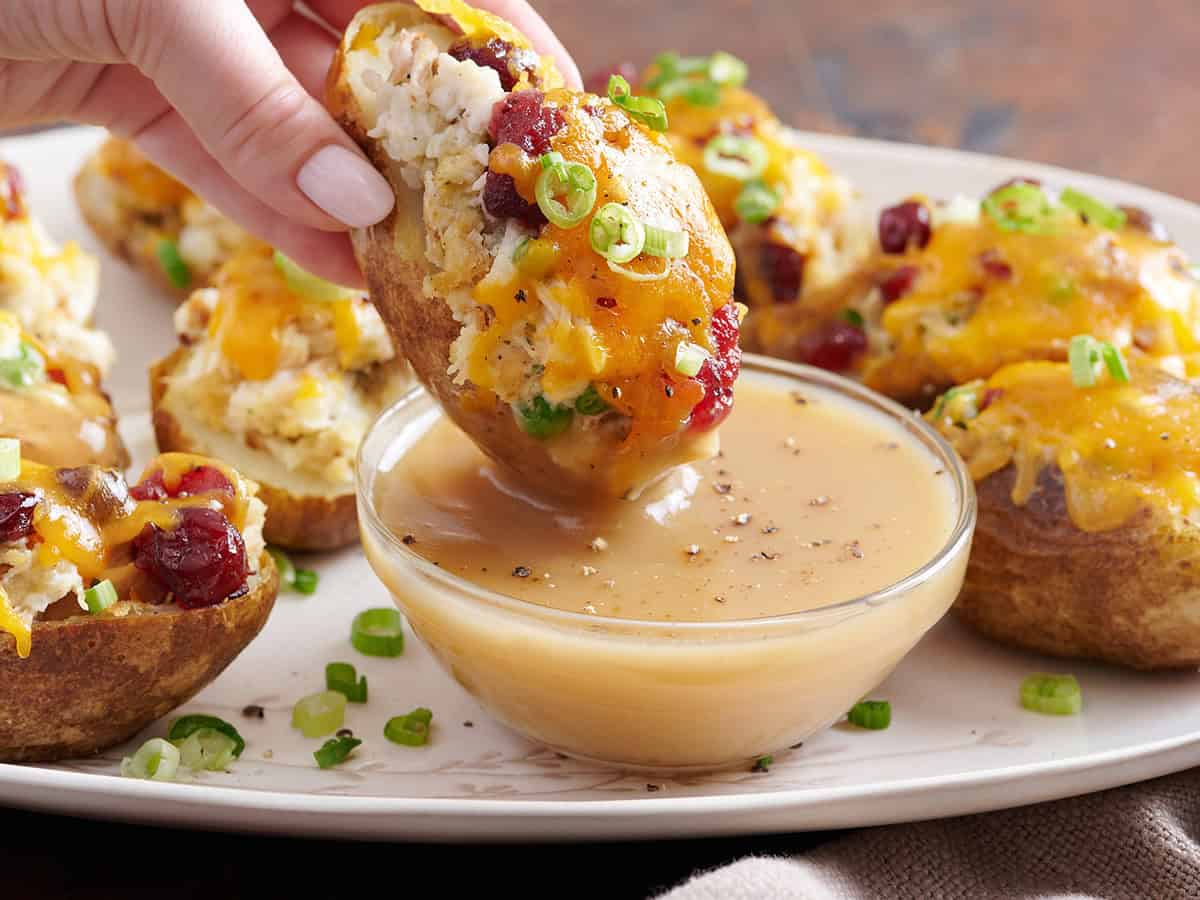 Side view of thanksgiving leftover potato skins on a plate, with a hand holding one and dipping it into a bowl of gravy.