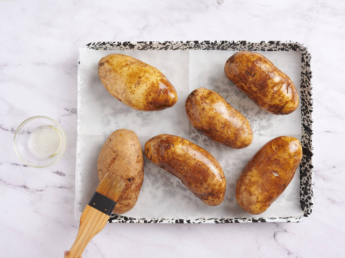 Overhead view of potatoes on a baking sheet with a brush brushing oil over the potatoes.