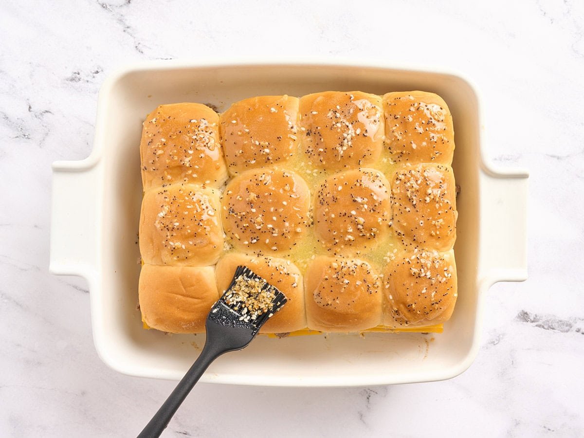 A pastry brush brushing seasoned melted butter over sliders in a casserole dish.