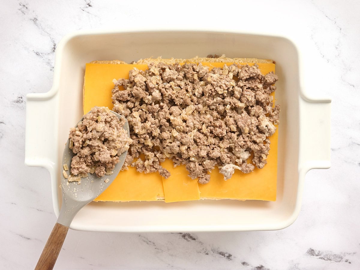 A spoon layering cooked ground beef onto cheese topped slider rolls in a casserole dish.