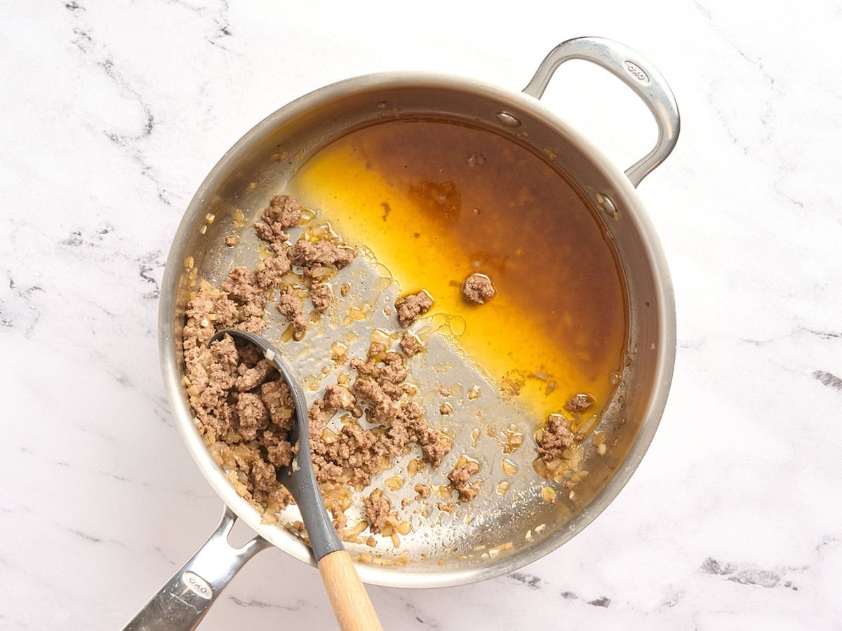 A spoon draining the fat from cooked ground beef in a skillet.