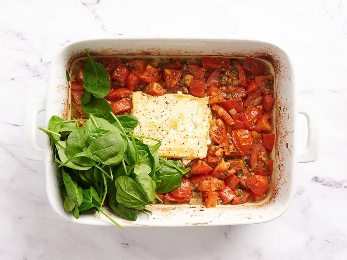 Spinach added to baked feta and tomatoes in a baking dish.