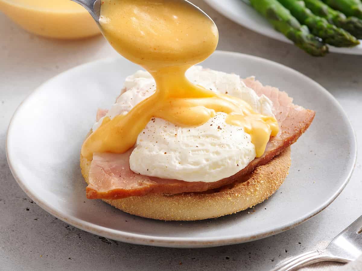 Homemade hollandaise sauce being drizzled onto eggs and ham.