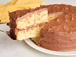 A homemade vanilla cake with chocolate frosting, with a slice being taken.