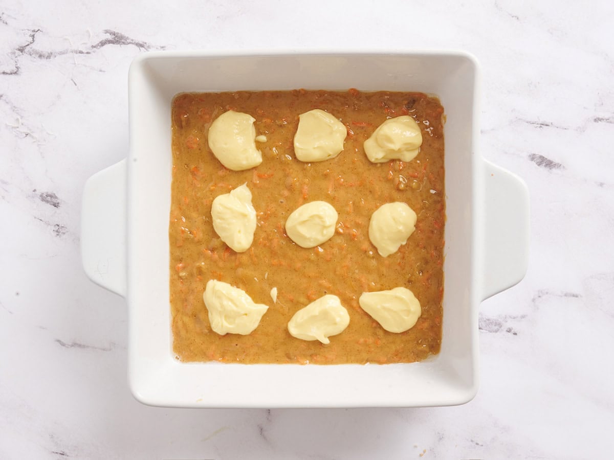 A layer of carrot cake bar mix added to a baking dish with cheese cake mixture dolloped on top.