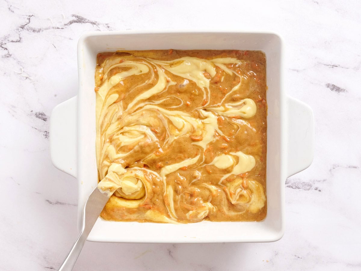 Cheesecake swirls on carrot cake bars in a baking dish.