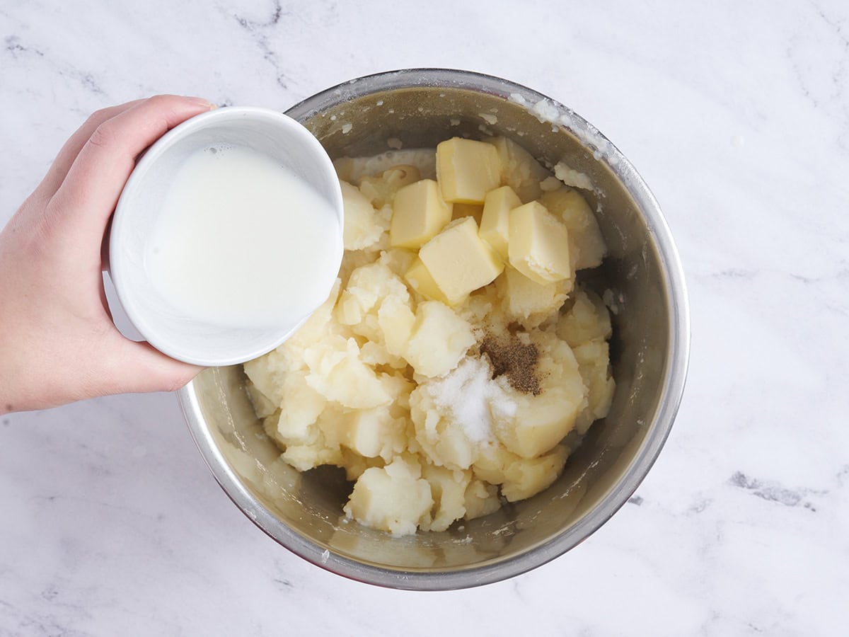 A hand pouring warm milk into an instant pot of cooked potatoes.
