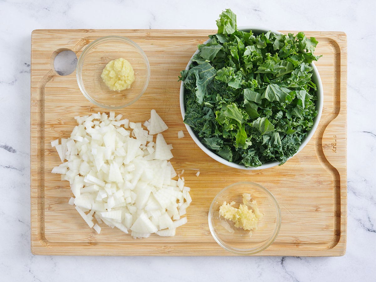 Diced onion, minced garlic and ginger, and chopped kale on a wooden cutting board.