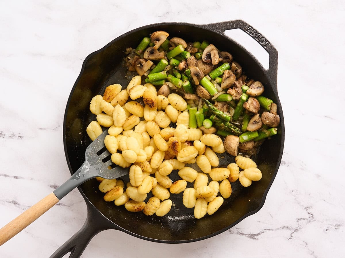 Gnocchi being flipped with a spatula in a skillet with asparagus and mushrooms.