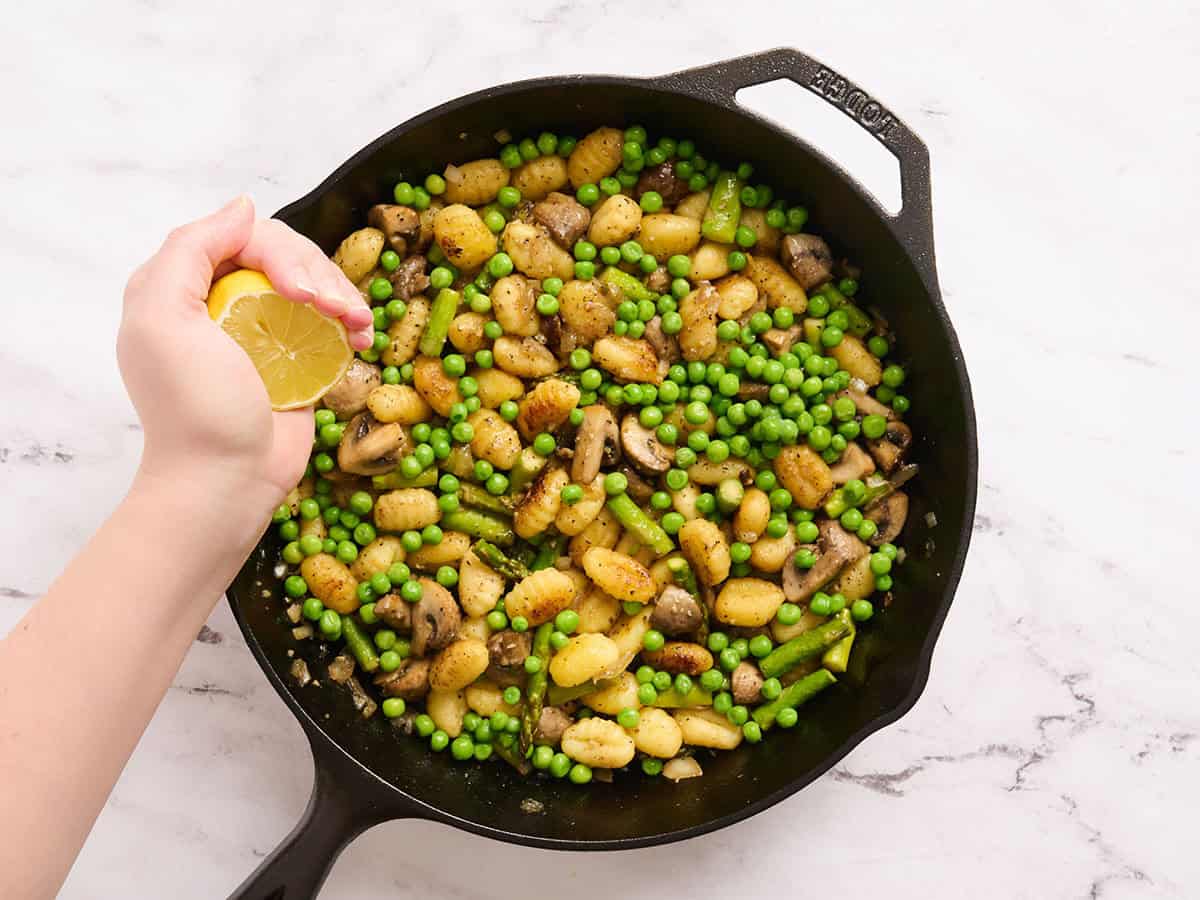 A lemon being squeezed by a hand into a pan of gnocchi with spring veggies.