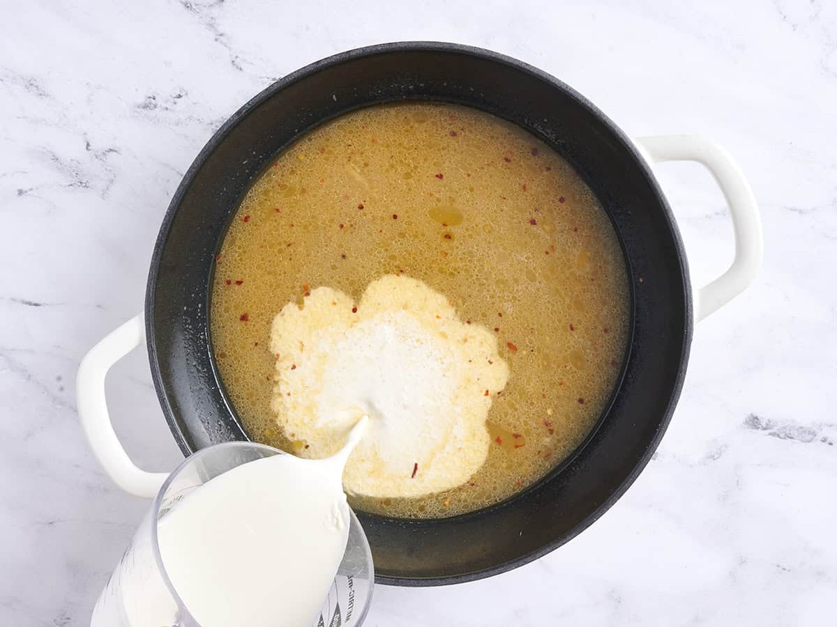 Heavy cream being poured into a pan with chicken broth.