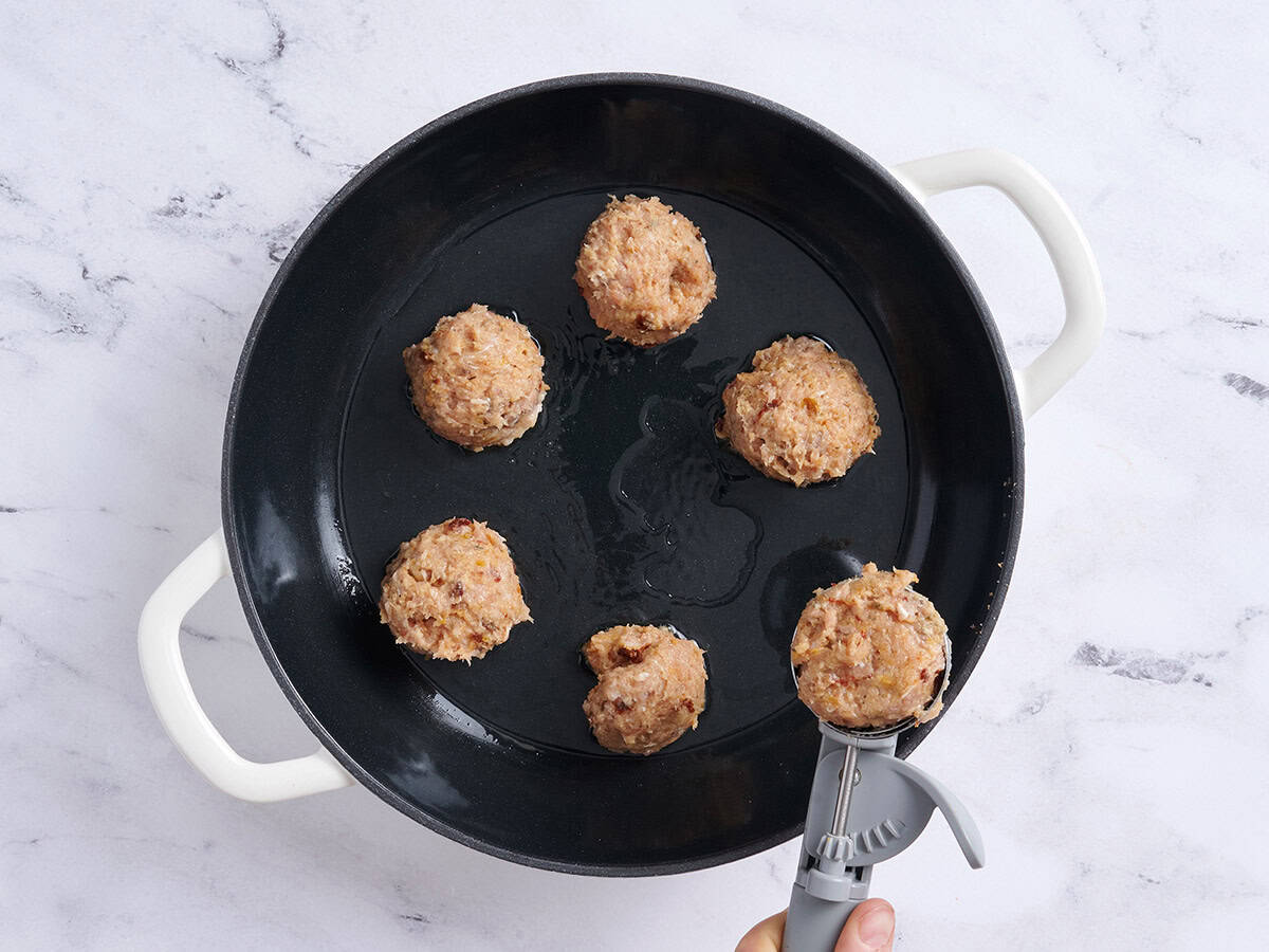 An ice cream scoop adding meatballs to a skillet.