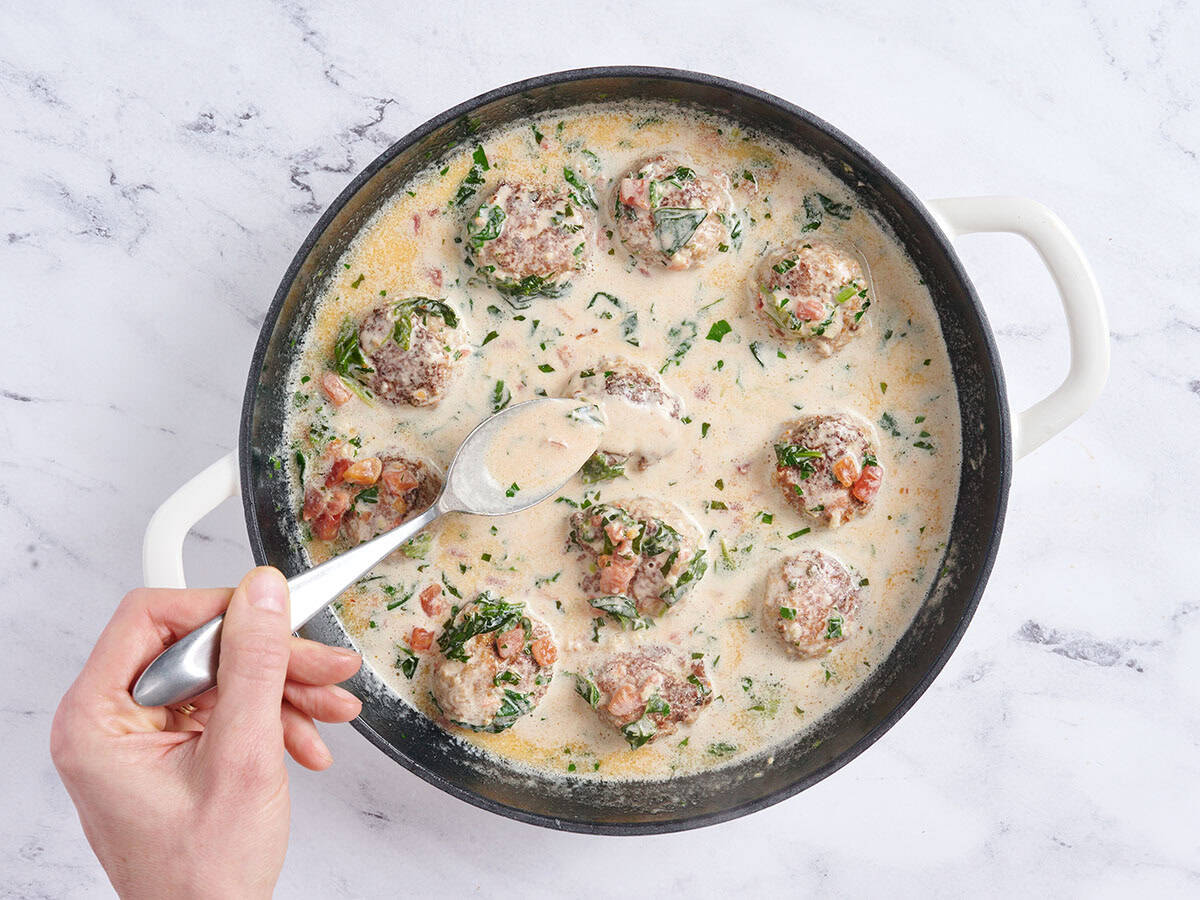 A hand using a spoon to braise chicken meatballs in a creamy sauce.
